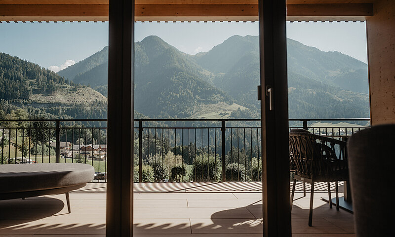 Balkon mit Sitzmöbeln und weiter Aussicht auf bewaldete Berge im Sonnenschein.