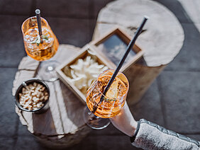 Zwei Gläser mit orangefarbenen Getränken stehen auf einem rustikalen Tisch, umgeben von Snacks wie Nüssen und Chips.