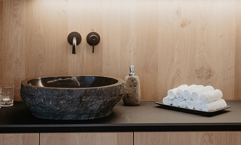 Stilvoll gestalteter Waschbereich im Wellnessbereich des Hotel Berghof mit Natursteinbecken, Handtüchern und Holzverkleidung.