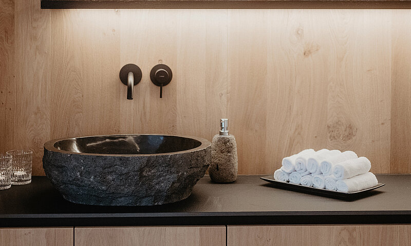 Stilvoll gestalteter Waschbereich im Wellnessbereich des Hotel Berghof mit Natursteinbecken, Handtüchern und Holzverkleidung.