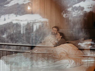 Paar liegt gemütlich im Bett in einem Zimmer; durch das Fenster spiegelt sich eine verschneite Berglandschaft.