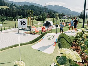 Eine Familie spielt Minigolf auf einem sonnigen Platz mit grünen Hügeln und blühenden Pflanzen im Hintergrund.
