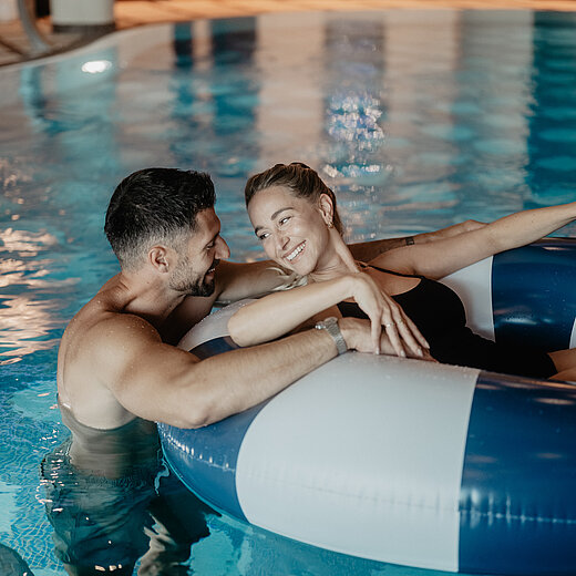Paar entspannt gemeinsam auf einem Schwimmring im Indoor-Pool des Hotel Berghof.