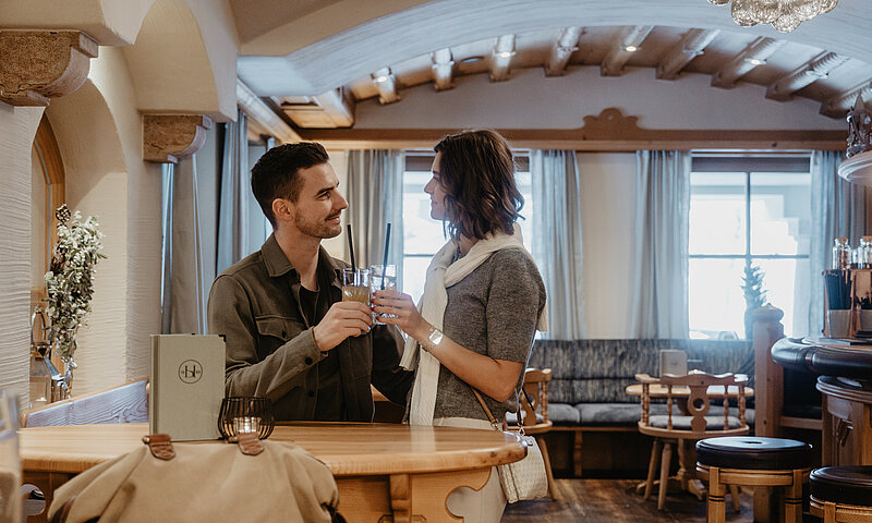 Ein Paar stößt mit Getränken an einem Stehtisch in einer gemütlichen Hotelbar im alpinen Stil an.