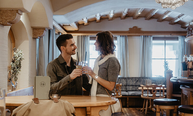 Ein Paar stößt mit Getränken an einem Stehtisch in einer gemütlichen Hotelbar im alpinen Stil an.