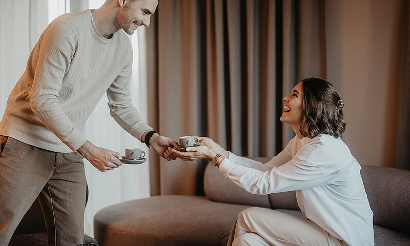 Ein Mann reicht seiner Freundin einen Kaffee, den er im Zimmer zubereitet hat.