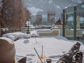 Eine Person entspannt im Schnee, genießt einen Cocktail und blickt auf die schneebedeckten Berge im Hintergrund.