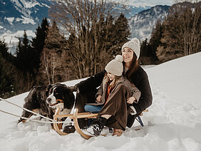Frau und Kind sitzen auf einem Schlitten im Schnee und nebenan sitzt ein Hund.