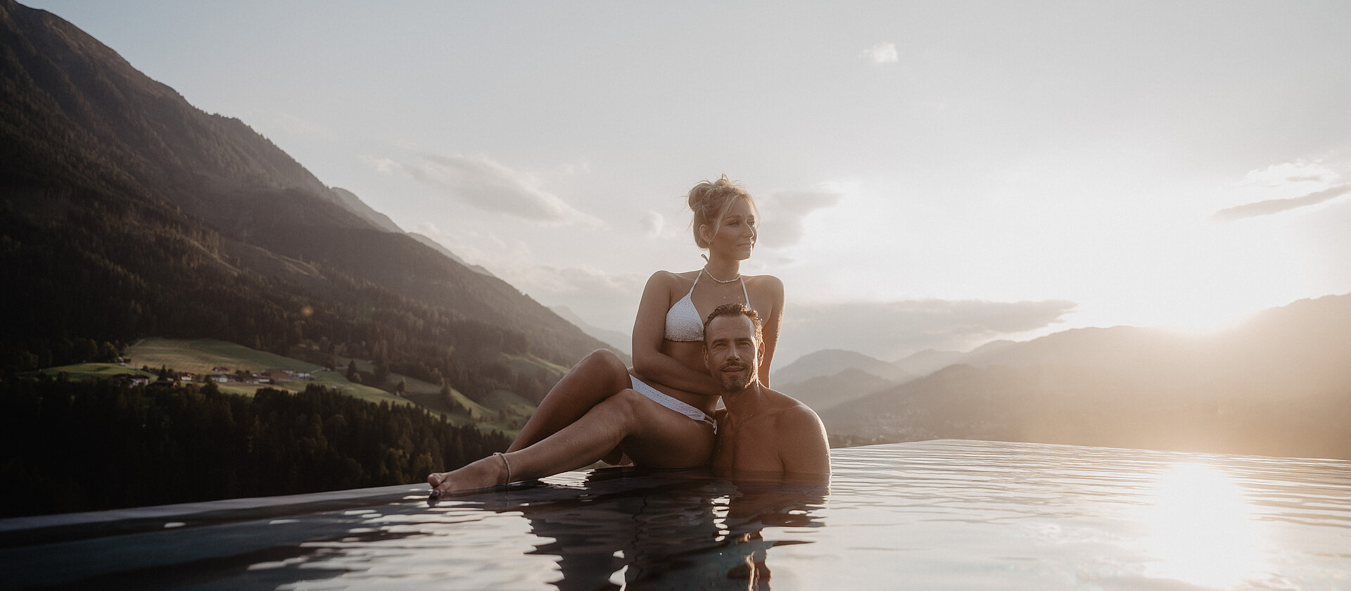 Paar entspannt im Infinitypool und genießt den Sonnenuntergang mit Bergpanorama im Hotel Berghof Salzburg