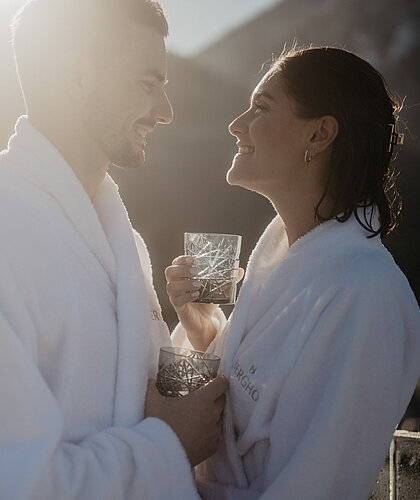 Paar in Bademänteln genießt einen Drink bei Sonnenlicht mit Bergblick auf der Terrasse Bergpanorama im Hotel Berghof.