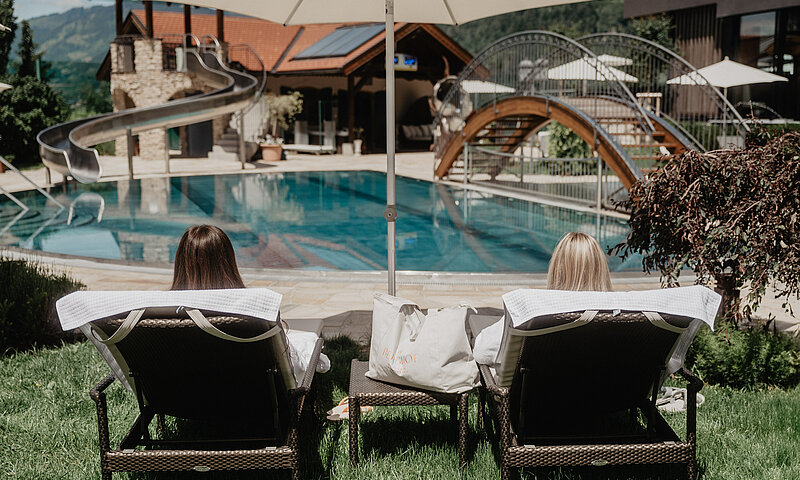 Zwei Frauen entspannen auf Liegen am Pool des Hotel Berghof Salzburg unter Sonnenschirmen.
