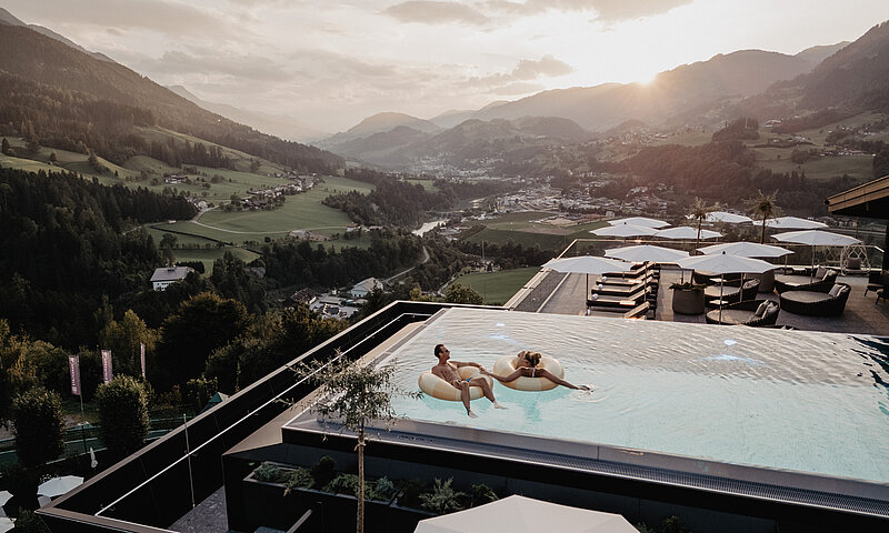 Zwei Personen werden aus der Vogelperspektive fotografiert wie sie auf Schwimmreifen im Infinity Pool des Hotel Berghof entspannen.