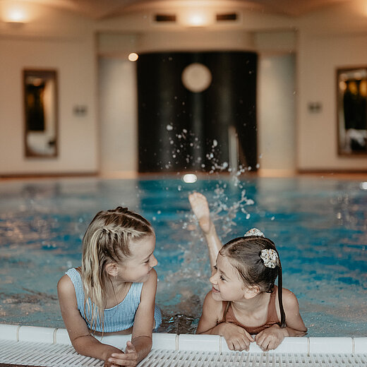 Zwei Mädchen strampeln im Indoor Pool des Hotel Berghofs und lachen sich dabei an.