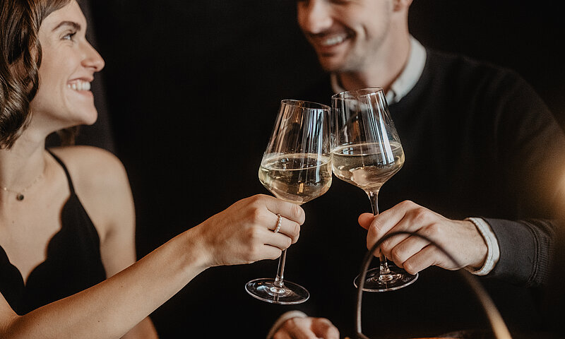 Zwei Personen die miteinander anstoßen mit einem Glas Wein und sich verliebt in die Augen schauen.