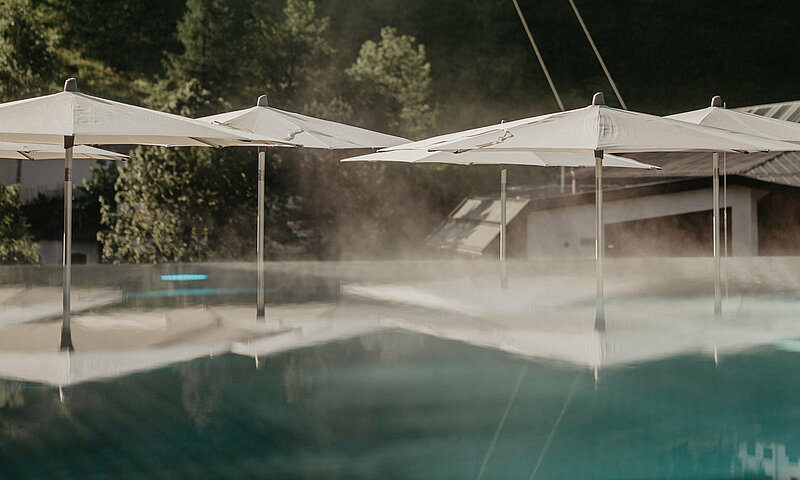 Beheizter Infinitypool mit aufsteigendem Wasserdampf und Sonnenschirmen im Wellnesshotel Berghof in Salzburg, eingebettet in grüne Berglandschaft.