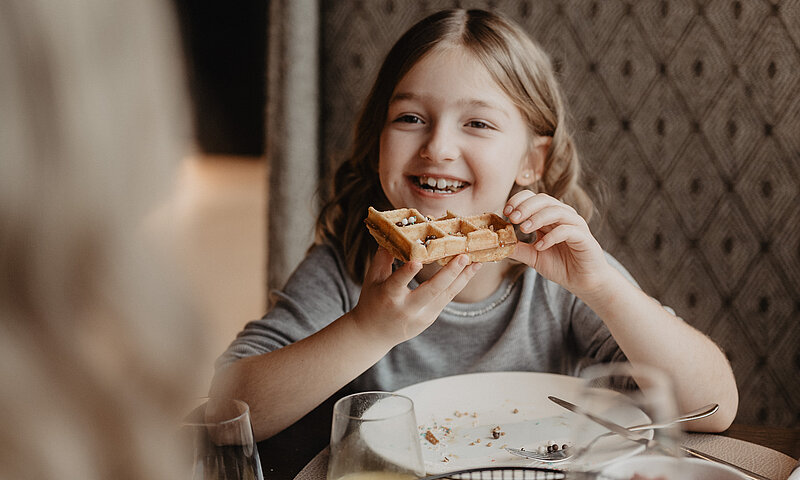 Kind lächelnd am Frühstückstisch mit frischer Waffel in der Hand.