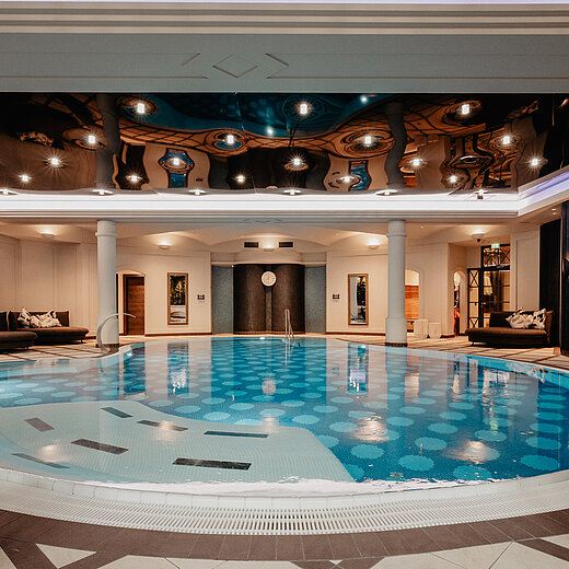 Indoor-Pool im Wellnessbereich des Hotel Berghof mit stilvollen Liegeflächen, weißen Säulen und einer spiegelnden Decke mit indirekter Beleuchtung.