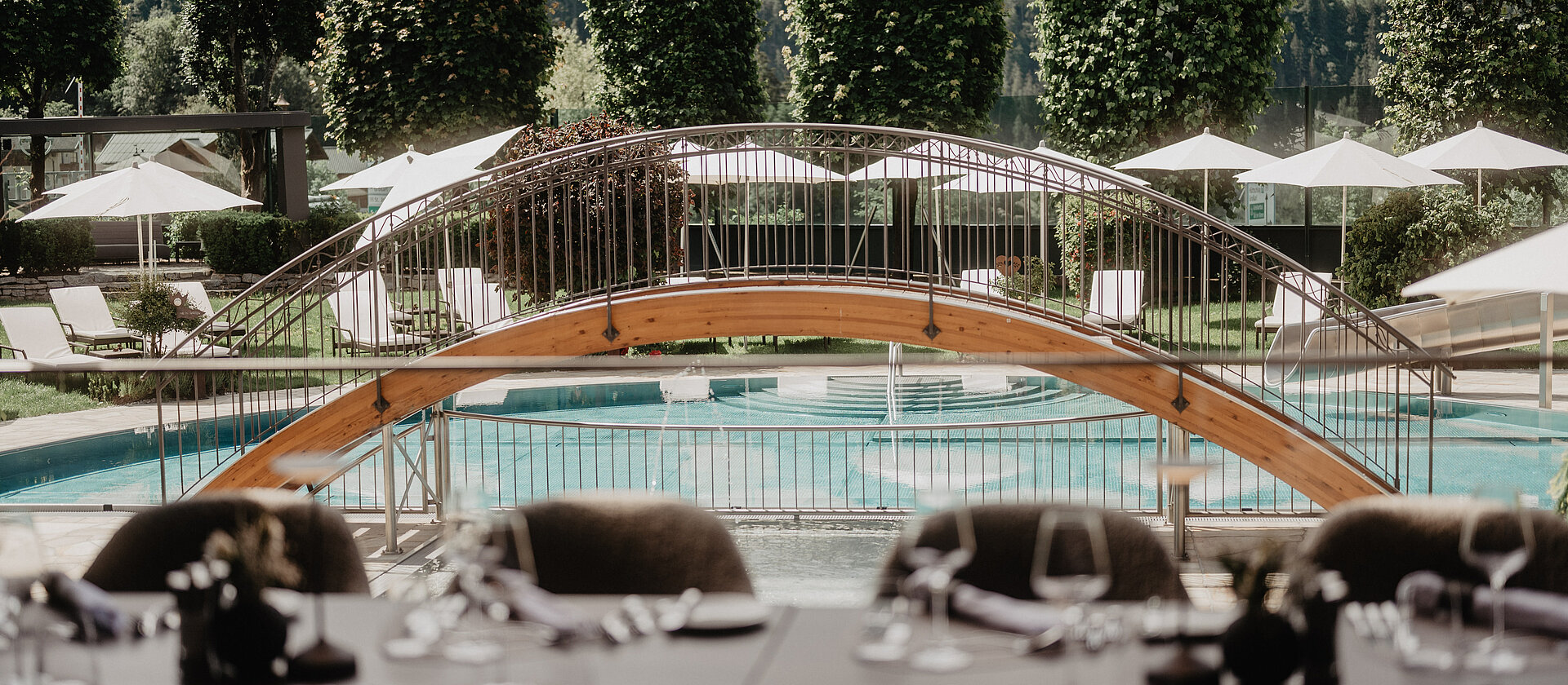 Blick von der Terrasse des Hotel Berghof auf den Außenpool mit kleiner Holzbrücke und gepflegtem Garten im Sommer.