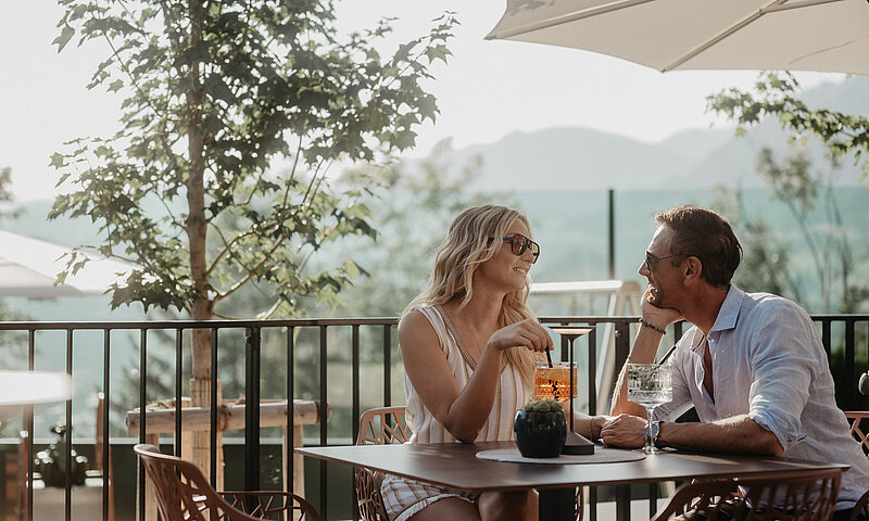 Ein Paar sitzt auf der Panoramaterrasse des Hotel Berghof und genießt Drinks im Sonnenschein.