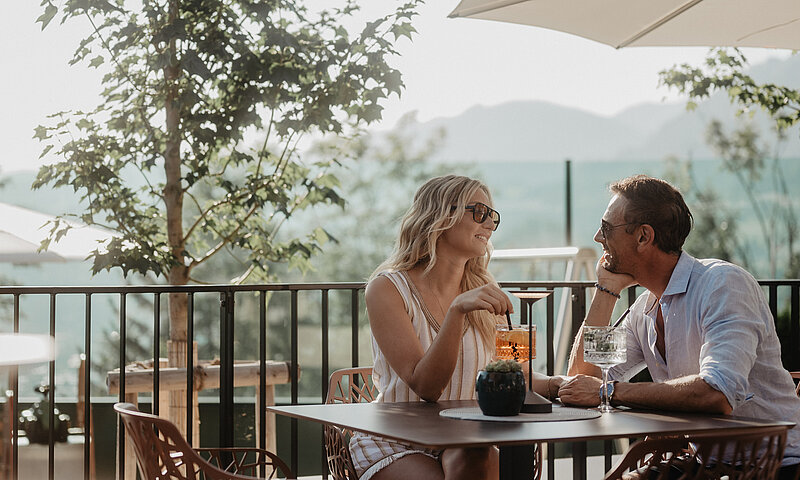 Ein Paar sitzt auf der Panoramaterrasse des Hotel Berghof und genießt Drinks im Sonnenschein.