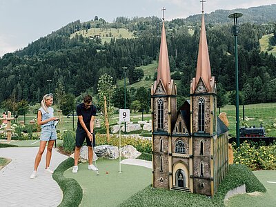 Ein Mann spielt Minigolf vor einer Miniaturkirche mit zwei Türmen, während eine Frau zuschaut und lächelt.