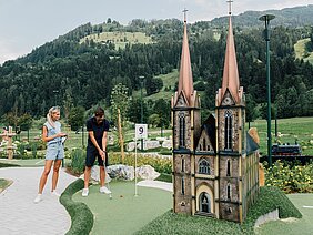 Ein Mann spielt Minigolf vor einer Miniaturkirche mit zwei Türmen, während eine Frau zuschaut und lächelt.