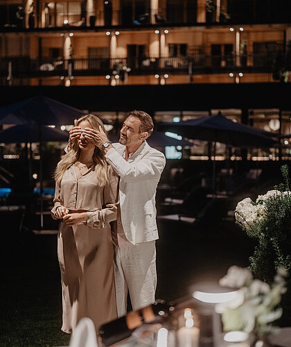 Ein Mann hält einer Frau spielerisch die Augen vor dem Tisch beim Abendessen zu.