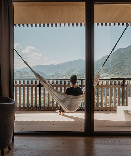 Eine Frau genießt die Aussicht auf die Berge, während sie in einer Hängematte auf dem Balkon der Panorama Suite im Wellnesshotel Salzburg sitzt und die Sonne scheint.