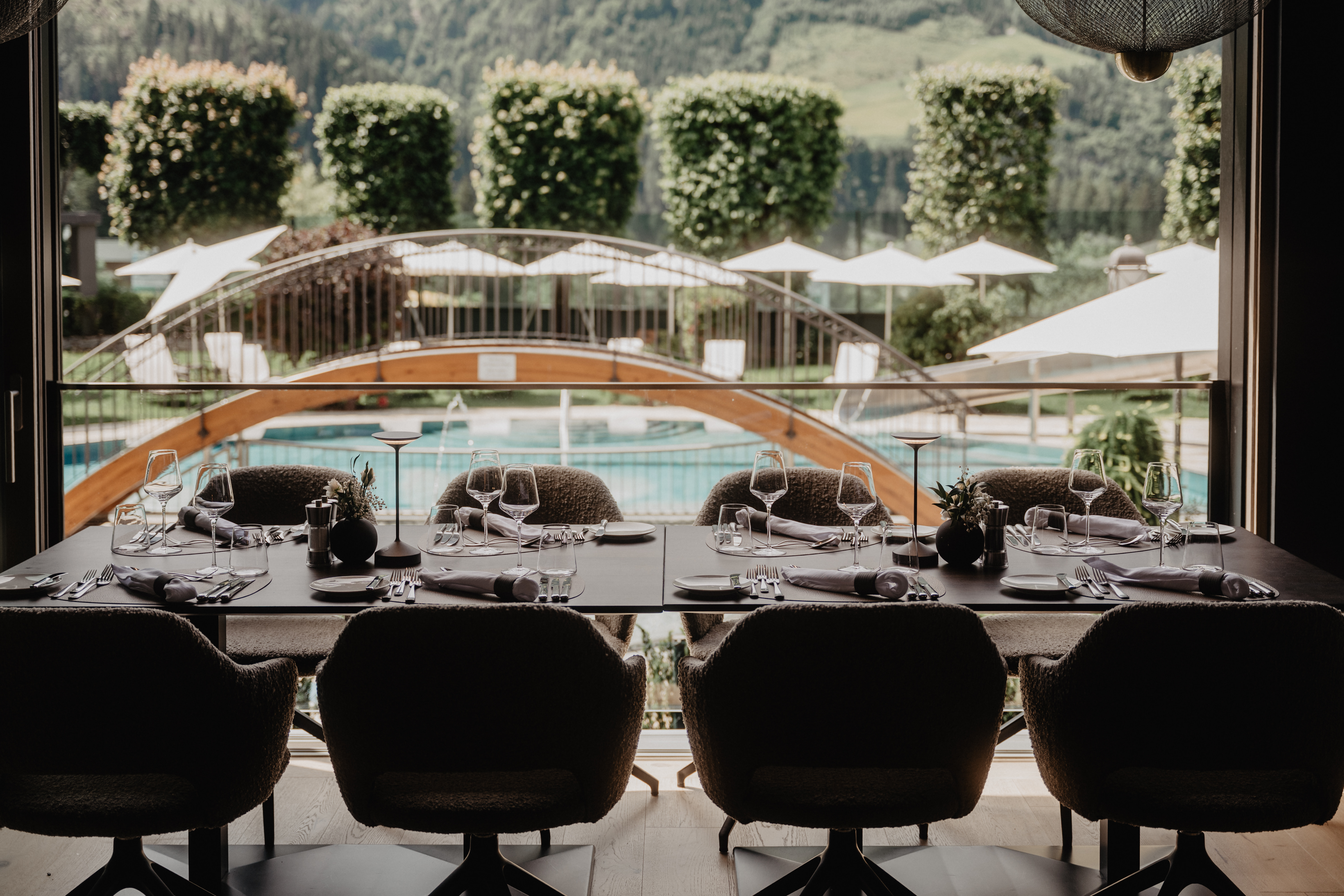 Blick von der Terrasse des Hotel Berghof auf den Außenpool mit kleiner Holzbrücke und gepflegtem Garten im Sommer.